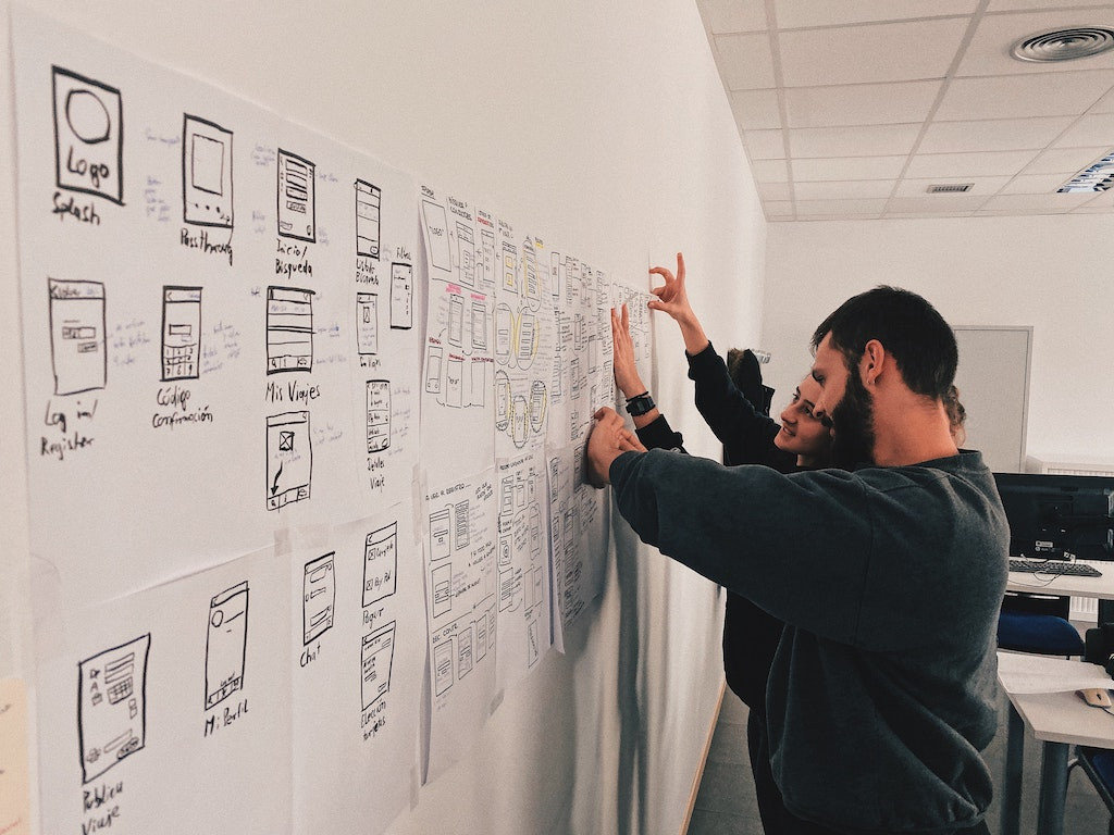 A diagonal side view of a whiteboard covered in website storyboards, with a bearded man and young woman standing at the far end, attaching new sheets to the board