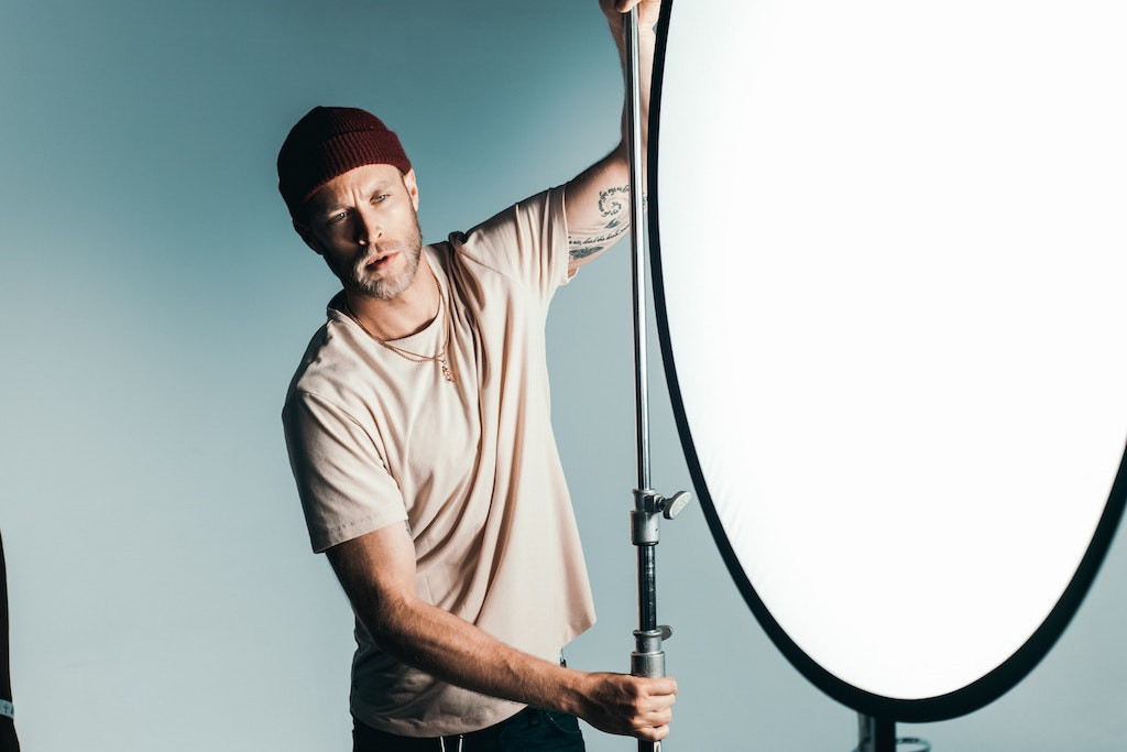 A hipster with a maroon skullcap folded above his ears, a beige t-shirt, subtle gold necklaces, centered, holding large production-quality lighting equipment for a LinkedIn video 