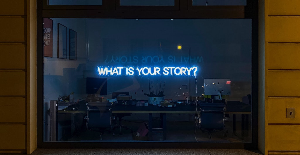 A neon sign reading 'WHAT IS YOUR STORY?' in the middle of a glass window, with an office seen through the glass