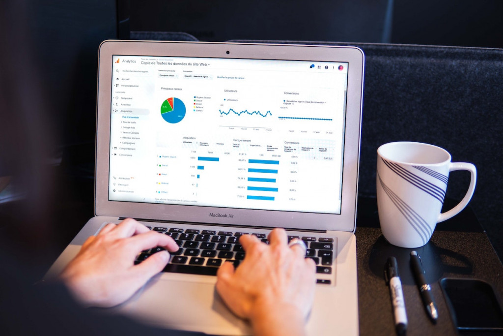 An open Macbook Air laptop, displaying Google Analytics on the screen, from the perspective of the user, with only hands showing; a white coffee mug with blue lines on it, as well as a Sharpie and pen, sit to the right of the computer