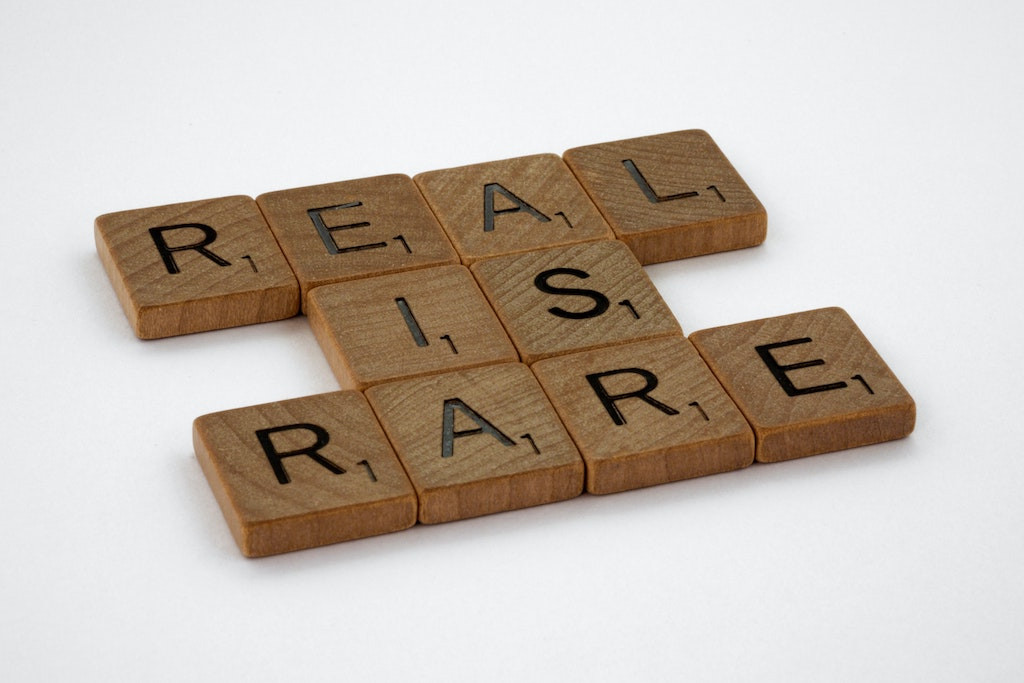 Over a white background, 10 traditional light brown wood Scrabble pieces arranged to read: (line 1) REAL (line 2) IS (line 3) RARE