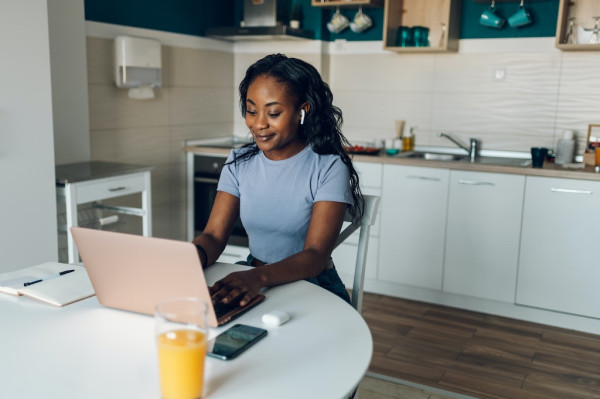 A Black woman with long, wavy black hair, a light blue short-sleeved shirt, and Apple AirPods in her ears, sits upright, smiling, at a white table, typing on her laptop, surrounded by her computer mouse, mobile phone, notebook, pen and glass of orange juice, as she works from home in her kitchen